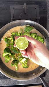 Hand holding half a lime over a simmering broccoli curry, ready to squeeze.