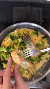 Close-up of a fork holding a flaky piece of cooked salmon over broccoli curry.