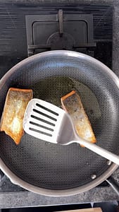 Two salmon fillets being seared skin-side down in a hot pan.